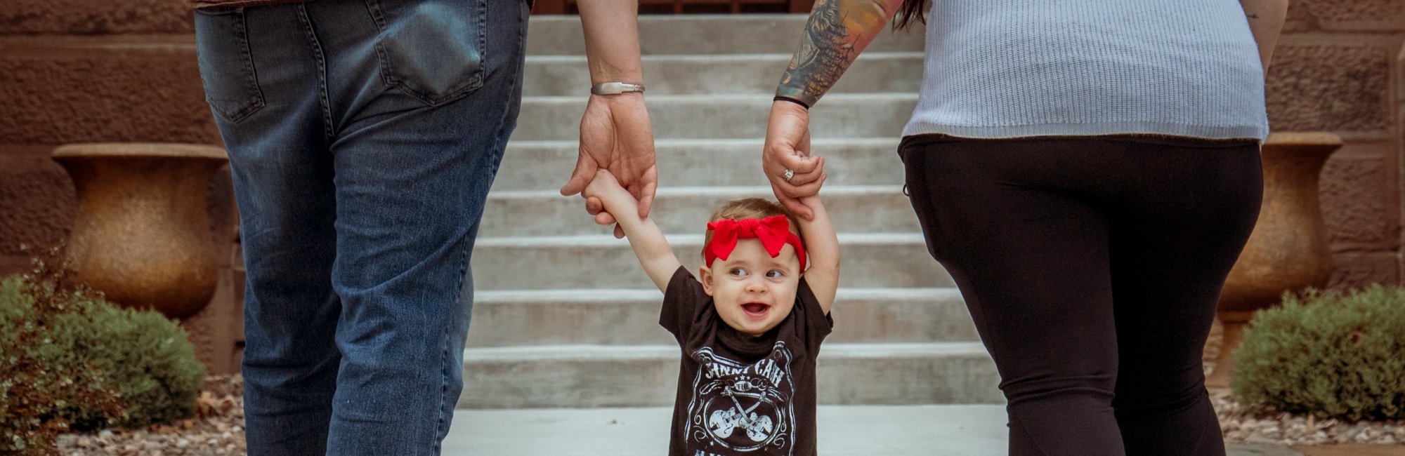 child with red bow in hair holds both parents hands