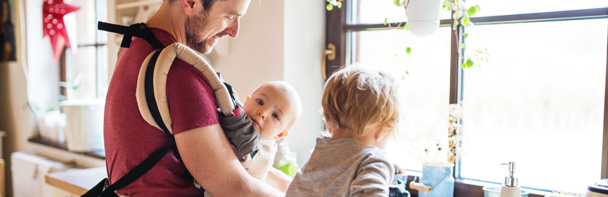 father wears baby in carrier in daylight in kitchen while other older child plays next to them