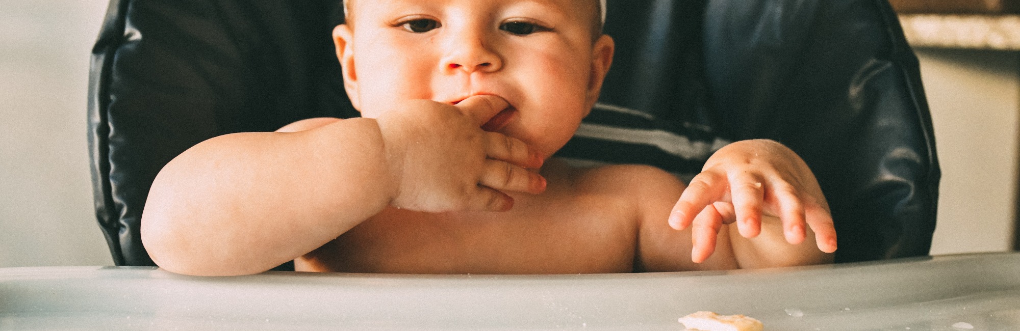 baby in highchair sucks on its own finger while playing with food