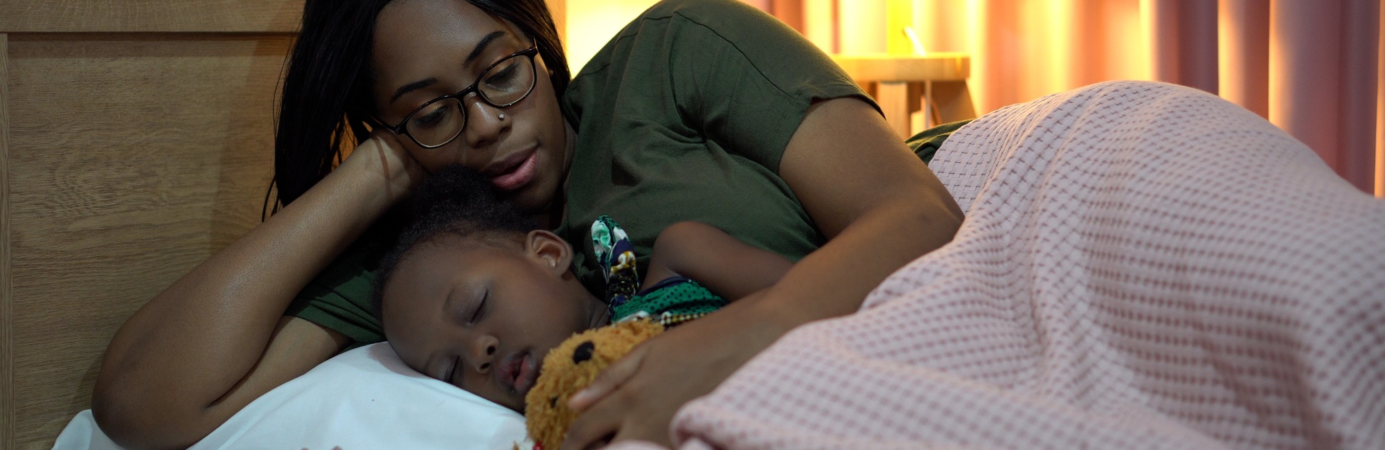 dark skinned woman with nose piercing holds her sleeping child as it sleeps in bed
